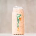 A clear glass bottle labeled Hummus FIT is filled with a thick, light orange smoothie. The background is neutral and out of focus, drawing attention to the bottle.