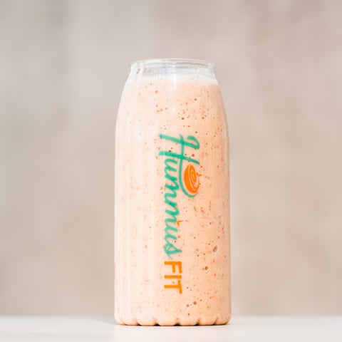 A clear glass bottle labeled Hummus FIT is filled with a thick, light orange smoothie. The background is neutral and out of focus, drawing attention to the bottle.