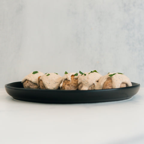 Keto Ricotta Meatballs—four meatballs with creamy sauce and fresh herbs on a black plate, set against a light background—make for a tasty, high-protein choice.