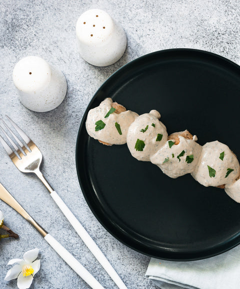 Four meatballs covered in creamy white sauce and garnished with herbs are arranged on a black plate. Nearby are a fork, knife, white salt and pepper shakers, a napkin, and a small flower on a light gray surface.