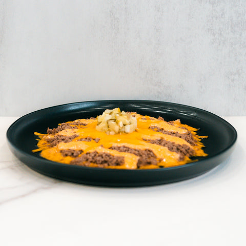 The Low Carb Keto Cheeseburger Bowl features ground beef topped with melted cheddar cheese in stripes and a center pile of diced pickles on a black plate—a high-protein, low-carb comfort dish against a minimalistic background.