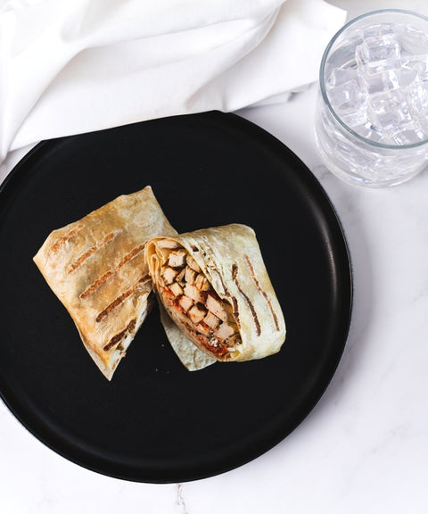 A grilled wrap cut in half, revealing grilled chicken and vegetables inside, sits on a black plate. Next to it is a glass of ice water on a white marble surface with a white napkin.