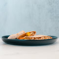 A blue plate with two HFit Signature Folds—filled with cheese, yellow bell peppers, and grilled chicken—sits on a white surface against a light blue textured background, making it a perfect option for healthy meal prep.