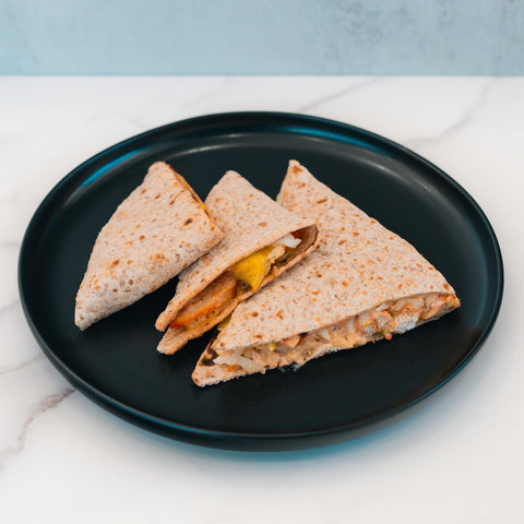 Three triangular HFit Signature Fold pieces with a whole wheat flatbread exterior and visible grilled chicken filling, arranged on a round black plate atop a white marble surface—a perfect option for healthy meal prep.