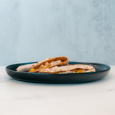 Two folded Strongsville Chicken Ranch Folds, filled with melted cheese and juicy chicken, are served on a dark blue plate atop a white marble surface with a light blue background. This high-protein meal is a deliciously satisfying option for any time of day.
