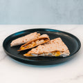 Two Strongsville Chicken Ranch Fold wedges with visible cheese and yellow bell peppers are served on a round black plate, set on a white marble surface with a light gray background—a delicious high-protein meal perfect for any time of day.