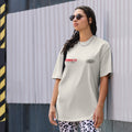 A woman wearing sunglasses and a light-colored T-shirt with the words HUMMUS FIT and EVOLVE stands against a metallic fence. She has braided hair and is also wearing patterned pants and a chain necklace.