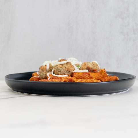 Rigatoni Meatballs—a black plate of rigatoni pasta in red sauce with ground-beef meatballs and melted cheese, set on a white surface with a light gray background—makes an easy, macro-friendly meal prep option.