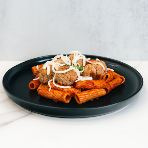 Rigatoni Meatballs: Rigatoni pasta in red sauce topped with three ground-beef meatballs and shredded cheese, served on a sleek black plate—ideal for meal prep or a macro-friendly dinner.