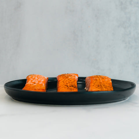 Three Competition Approved Lemon Pepper Salmon fillets 1lb are neatly arranged on a black plate, ideal for a low-carb, high-protein meal prep against a light, neutral background.