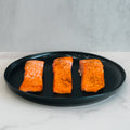 A 1lb pack of Competition Approved Lemon Pepper Salmon is displayed on a black plate atop a white marble surface with a light gray background—ideal for high-protein meal prep or low-carb diets.