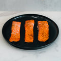 Three cooked Competition Approved Lemon Pepper Salmon fillets, lightly seasoned and arranged on a black plate, offer a delicious low-carb, high-protein meal prep option on a white marble surface.