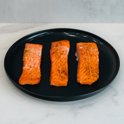 Three cooked Competition Approved Lemon Pepper Salmon fillets, lightly seasoned and arranged on a black plate, offer a delicious low-carb, high-protein meal prep option on a white marble surface.