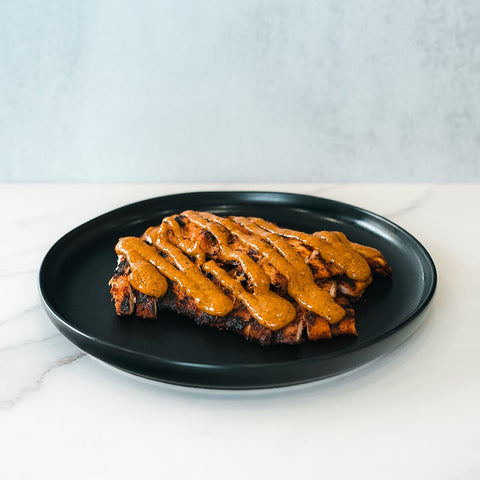 Competition Approved Grilled Chicken W/ Smokin Poppie Sauce 1lb is sliced and served on a black plate, topped with creamy orange sauce—an ideal high-protein meal displayed on white marble with a light gray background.