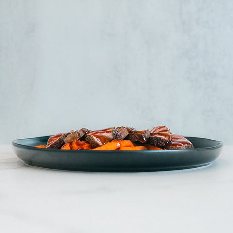 The Soho Steak Bowl features sliced steak with barbecue sauce over mashed sweet potatoes, served on a dark plate for a healthy, Asian-inspired lunch—presented on a white surface against a light gray background.