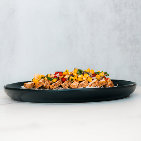 The Southwest Chicken Bowl features sliced grilled chicken, corn, diced red peppers, and herbs over white rice—a gluten-free meal prep option—served on a black plate with a light background.