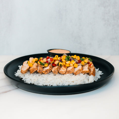 The Southwest Chicken Bowl features sliced grilled chicken with corn salsa over white rice on a black plate—gluten-free and meal-prep friendly. A small dipping sauce dish is in the background.