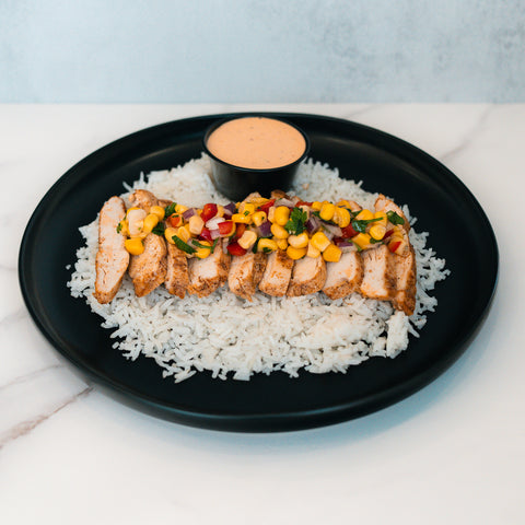 The Southwest Chicken Bowl is a gluten-free meal with sliced grilled chicken breast over white rice, topped with corn, diced tomatoes, and herbs. It comes meal-prep style on a black plate with a side of creamy orange sauce.