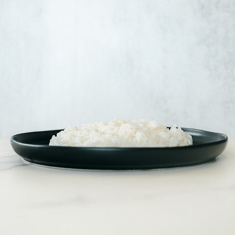 Competition Approved Sticky Rice 1lb—perfect for meal prep and packed with healthy carbs—served as a mound of white rice on a round black plate, set atop light marble with a simple, light background.