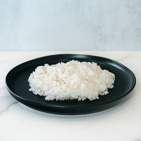 A round black plate holds Competition Approved Sticky Rice 1lb on a white marble surface against a light background—a simple, healthy carb option for meal prep.