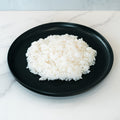 A mound of Competition Approved Sticky Rice 1lb is served on a round black plate atop white marble—ideal for meal prep and packed with healthy carbs.