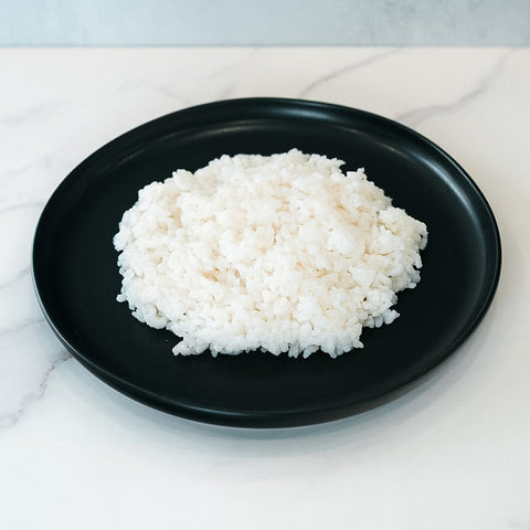 A mound of Competition Approved Sticky Rice 1lb is served on a round black plate atop white marble—ideal for meal prep and packed with healthy carbs.