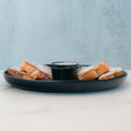 Six pieces of Blueberry French Toast are arranged in a circle on a black plate with a small cup of syrup in the center. Ideal for anyone craving a delicious breakfast, the plate rests on a white surface against a light blue, textured background.