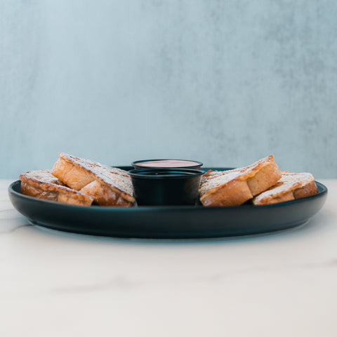 Six pieces of Blueberry French Toast are arranged in a circle on a black plate with a small cup of syrup in the center. Ideal for anyone craving a delicious breakfast, the plate rests on a white surface against a light blue, textured background.