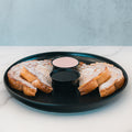 Slices of Blueberry French Toast are dusted with powdered sugar and arranged on a black plate, accompanied by two small cups of dipping sauces in the center. Served on a white marble surface, this delicious breakfast is as visually appealing as it is satisfying.