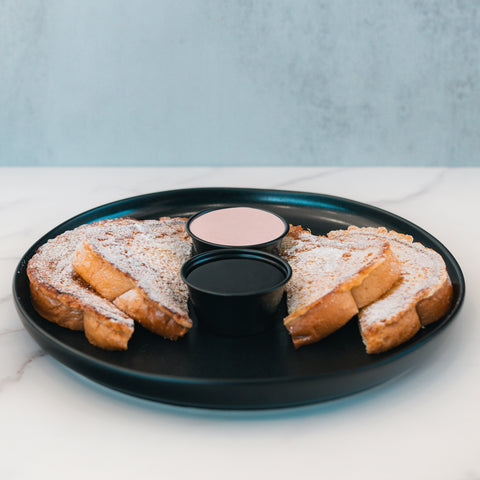 Slices of Blueberry French Toast are dusted with powdered sugar and arranged on a black plate, accompanied by two small cups of dipping sauces in the center. Served on a white marble surface, this delicious breakfast is as visually appealing as it is satisfying.