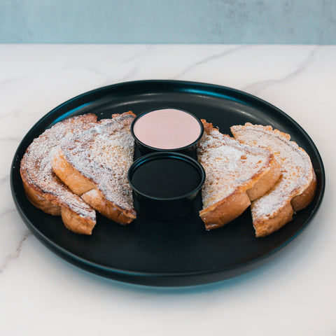 Four slices of Blueberry French Toast, dusted with powdered sugar, are arranged on a black plate alongside two small cups of dipping sauces—one pink and one dark—set on a white marble surface, making for a delicious high protein breakfast option.