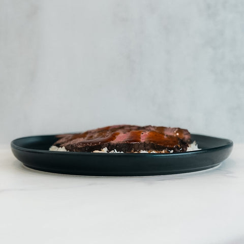 The Teriyaki Flank Bowl highlights glazed meat on fluffy white rice atop a dark green plate, styled against a minimalist light background—ideal for healthy eating and simple meal prep.
