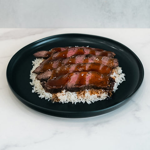 The Teriyaki Flank Bowl features sliced medium-rare steak with brown sauce over white rice, served on a black dish—a perfect choice for healthy meals and easy meal prep.