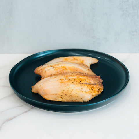 Three Baked Herbed Tilapia 1lb Competition Approved fillets are arranged side by side on a dark blue plate atop white marble—a delicious, healthy choice ideal for meal prep and weight loss goals.