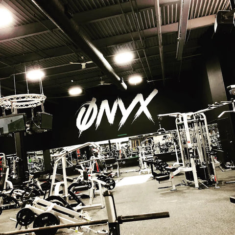 A modern gym with various fitness equipment and weight machines, featuring a large black wall with the word ONYX painted in bold, white, stylized letters. The gym appears spacious and well-lit.