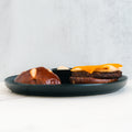 On a dark plate sits the Vegan Breakfast Sandwich: a split pretzel bun with a high-protein vegan patty, melted cheese, savory sauce, and a small cup of dipping sauce—presented on a white surface against a light gray background.