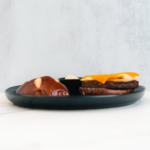 On a dark plate sits the Vegan Breakfast Sandwich: a split pretzel bun with a high-protein vegan patty, melted cheese, savory sauce, and a small cup of dipping sauce—presented on a white surface against a light gray background.