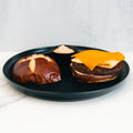 A deconstructed Vegan Breakfast Sandwich on a black plate, featuring a pretzel bun, plant-based patty topped with vegan cheddar and egg alternative, and a small cup of dipping sauce in the background.