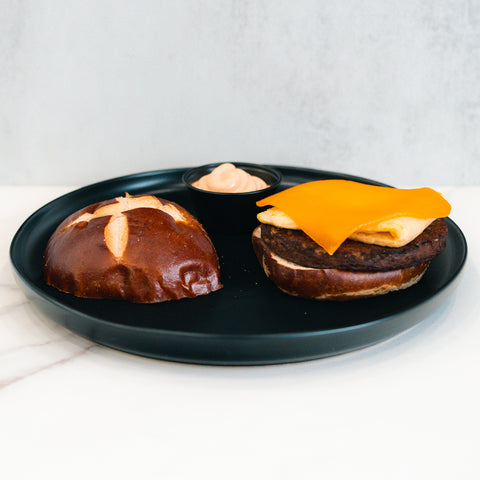 A deconstructed Vegan Breakfast Sandwich on a black plate, featuring a pretzel bun, plant-based patty topped with vegan cheddar and egg alternative, and a small cup of dipping sauce in the background.