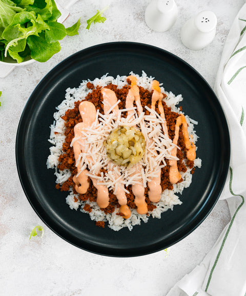 A black plate with white rice topped with seasoned ground meat, shredded cheese, creamy sauce, and a dollop of chopped pickles in the center. Fresh greens and salt and pepper shakers are nearby.