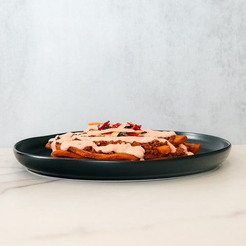 The Nacho Average Vegan Bowl features loaded fries with plant-based protein, pink creamy sauce, and red pepper bits, all served on a black plate atop white marble against a light gray background.