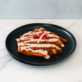 The Nacho Average Vegan Bowl features fries topped with plant-based protein, creamy pink sauce, and thinly sliced red and white veggies, all served on a black plate atop a white marble surface.