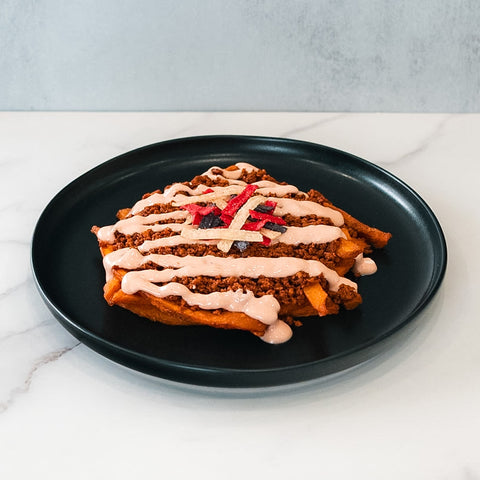 The Nacho Average Vegan Bowl features fries topped with plant-based protein, creamy pink sauce, and thinly sliced red and white veggies, all served on a black plate atop a white marble surface.