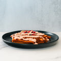 The Nacho Average Vegan Bowl features tamales with ground plant-based protein, pink sauce, and veggies on a black plate atop white marble—a perfect meatless option against a light grey background.