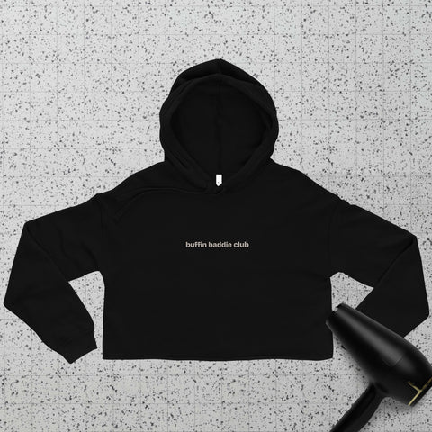 A black cropped hoodie with the text buffin baddie club in small white letters on the chest, laid flat on a speckled surface next to a black hair dryer.