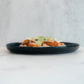 A Zeus Bowl sits on a black plate with green sauce and shredded cheese over Zeus chicken, beans, and jasmine rice, placed on a white surface against a light background.