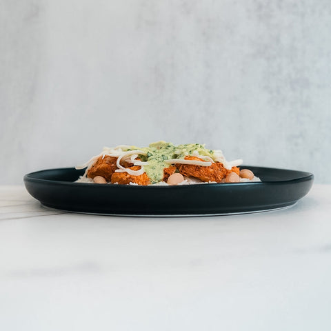 A Zeus Bowl sits on a black plate with green sauce and shredded cheese over Zeus chicken, beans, and jasmine rice, placed on a white surface against a light background.