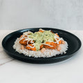 The Zeus Bowl features jasmine rice and white beans, topped with breaded Zeus chicken cutlets, shredded cheese, and creamy green herb sauce. Served on a black plate, it's presented on a white marble surface.
