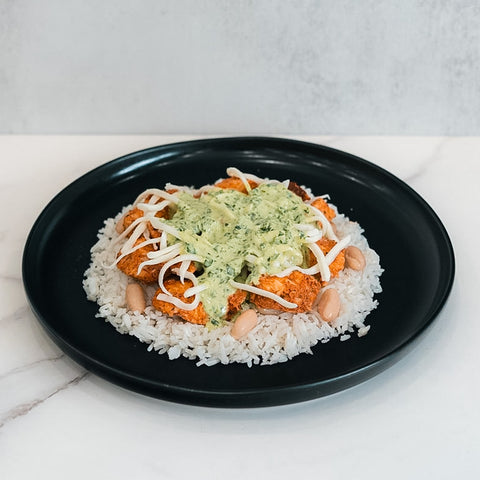 The Zeus Bowl features jasmine rice, beans, crispy Zeus chicken, beansprouts, and a creamy green herb sauce, served on a black plate. It is presented on a white surface against a light gray background.