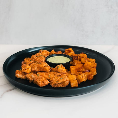 A black plate displays seasoned chicken kebab pieces and roasted potato cubes, with creamy green sauce in the center. Set on a light background, this presentation highlights the Mediterranean-inspired Zeus Bowl V2.